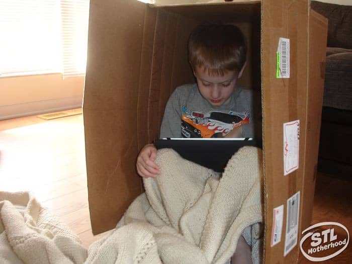 small boy sitting inside cardboard box with an ipad and blanket