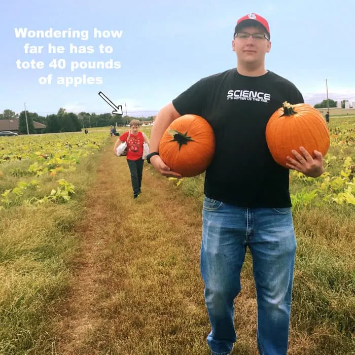 brothers carry fruit at Eckert's farm--teen is holding two pumpkins, one in each hand, smaller boy struggles to carry two bags of apples.