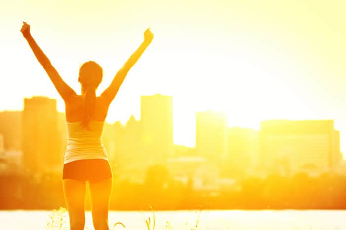 Success winner woman standing with arms up joyful after outdoors workout. Half silhouette on sunny warm summer day with city skyline in background,