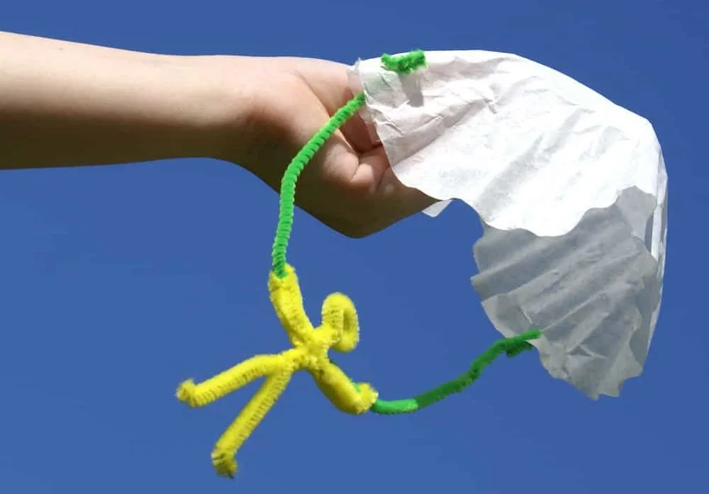 parachute man made of coffee filter and fuzzy sticks, held by child's hand against a blue sky