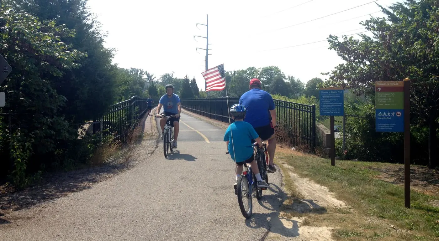 Dad and son on tagalong bike riding paved trail, with American flag on back of bike.