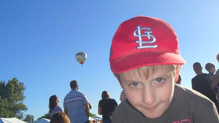 kid in red Cardinals hat smirks at camera with hot air balloons taking off behind him