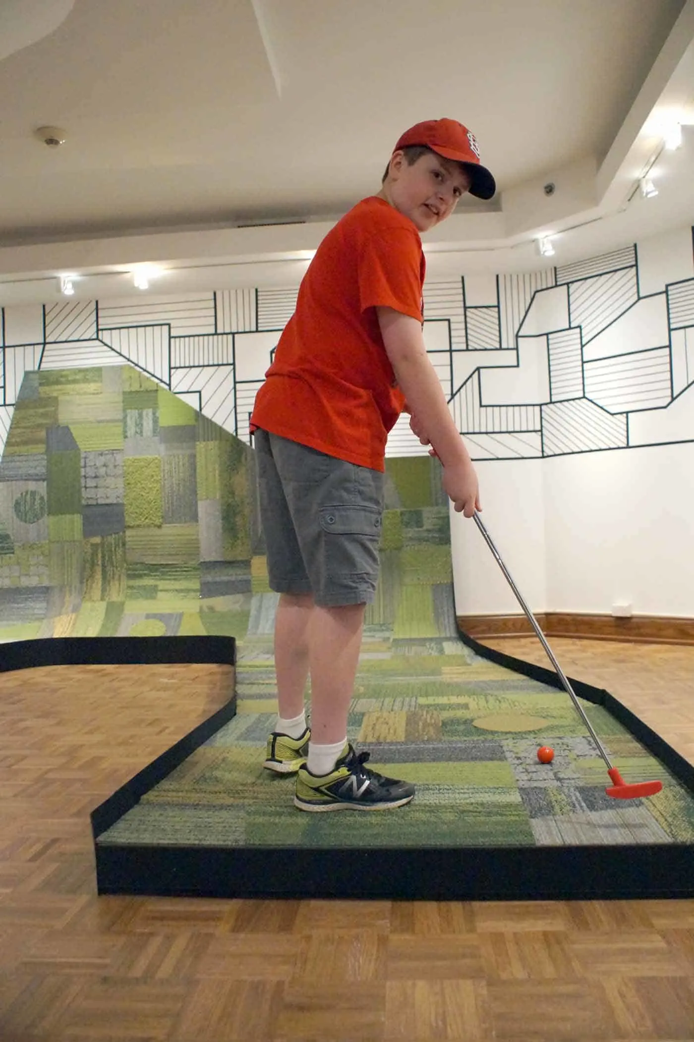 Indoor golf at an art museum. Boy in red shirt plays a mini golf hole made of green carpet squares running up the side of a wall.