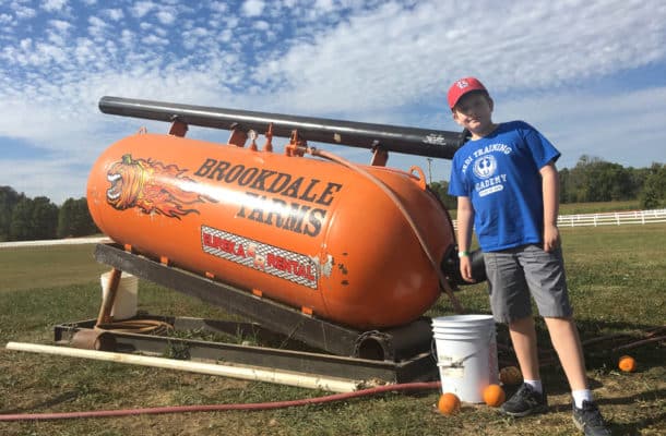 Kid in blue shirt by a giant air compressor powered pumpkin cannon.