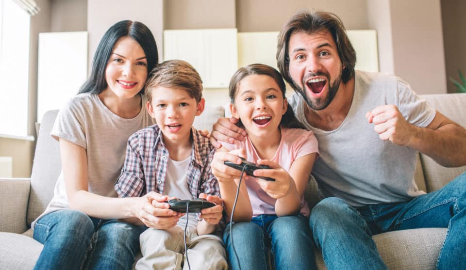 Amazing family sits on sofa. Children are playing on xbox. The game is intense. Guy rejoys. Girl is smiling as well. Mother helps to play her son