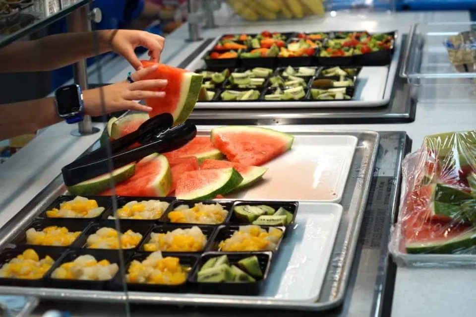 Child selecting a piece of bright red watermelon from a cafeteria food line