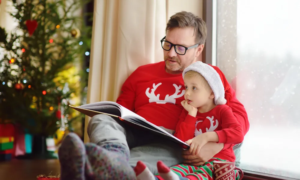 Man and child reading a book by a Christmas tree. Kid is wearing holiday pajamas. 