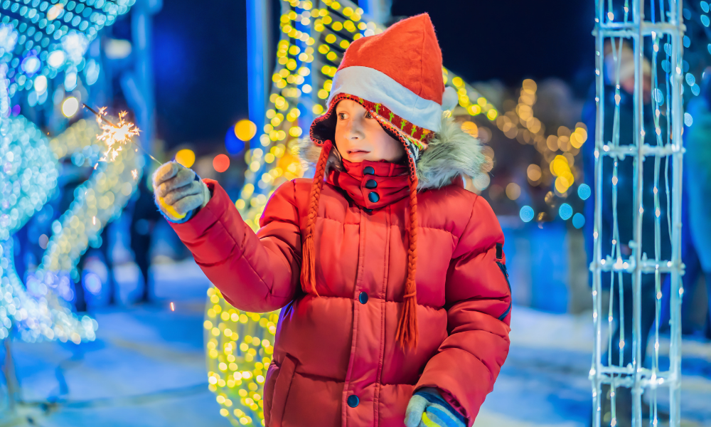 Kid in winter coat with twinkle lights, holding a sparkler.