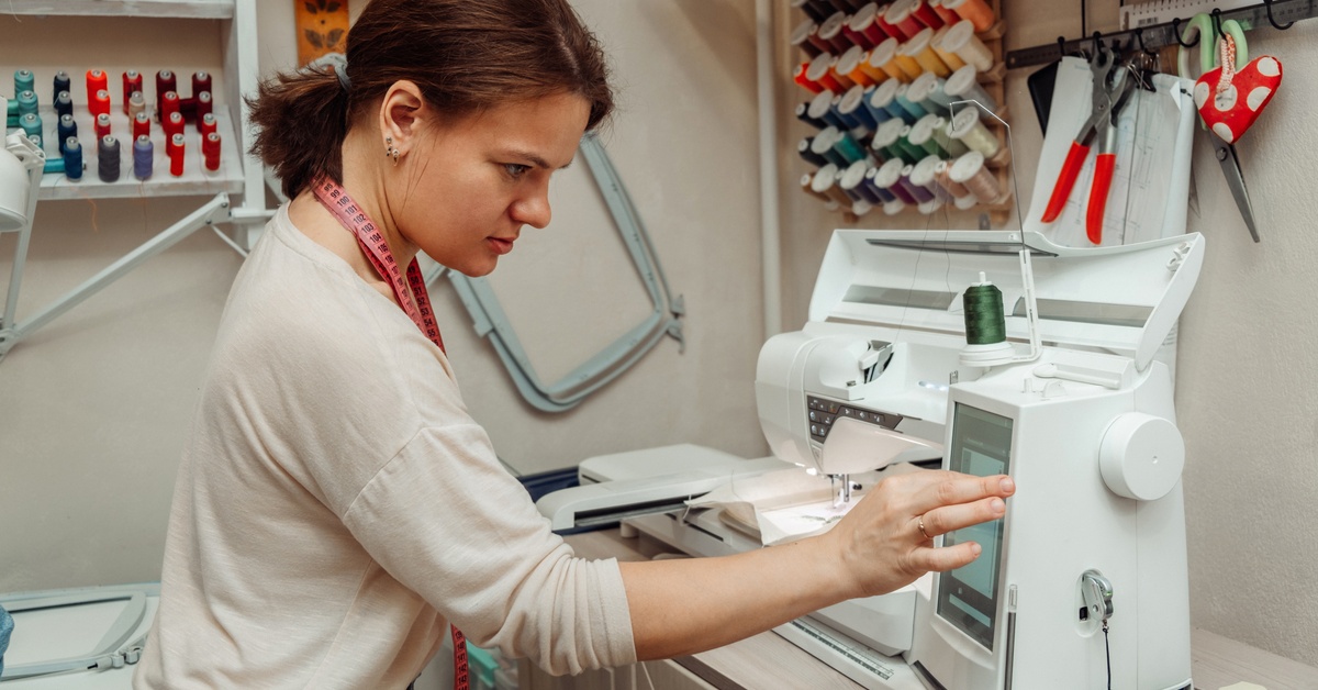 A woman operates an embroidery machine in a craft room, surrounded by colorful thread spools and sewing tools on the wall.