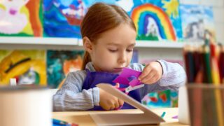 A young girl using safety scissors to cut a piece of purple paper. She is sitting in front of multiple art projects.