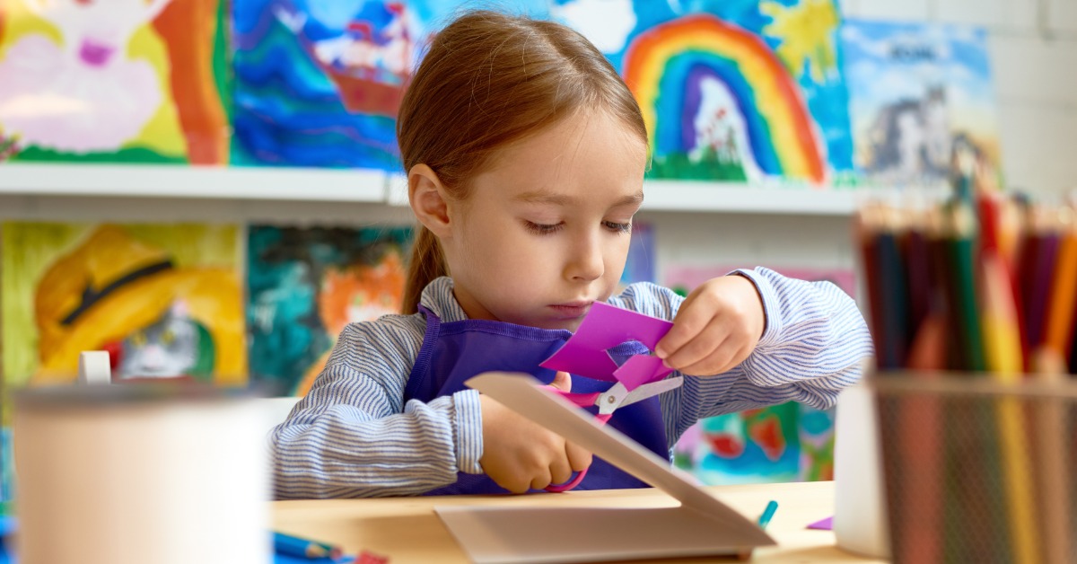 A young girl using safety scissors to cut a piece of purple paper. She is sitting in front of multiple art projects.