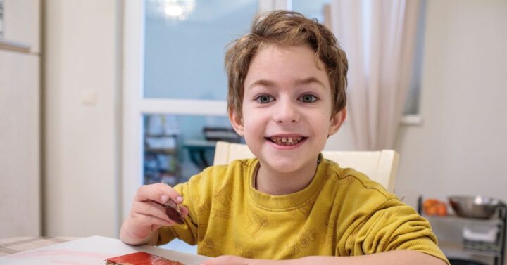 A little boy smiles as he paints a piece of cardboard with a brush. He sits at a table with a white covering over it.