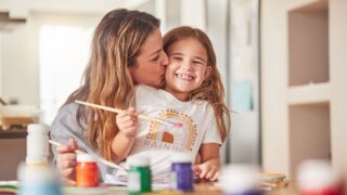 A woman is sitting behind her daughter and giving her a kiss on the cheek. The daughter is holding a paint brush.