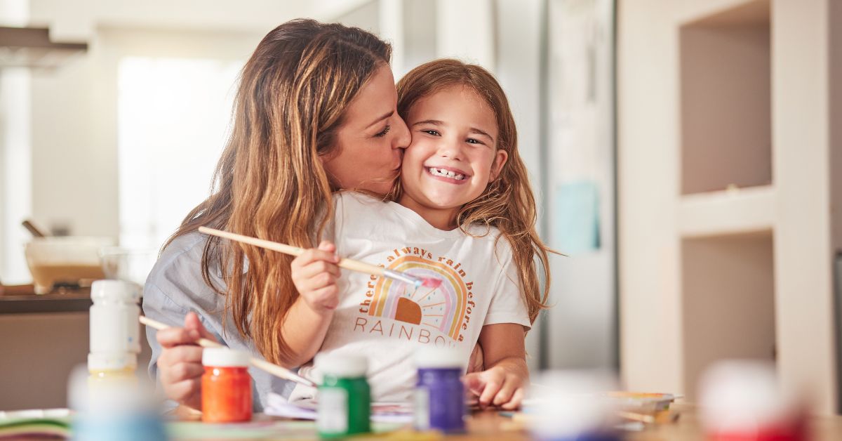 A woman is sitting behind her daughter and giving her a kiss on the cheek. The daughter is holding a paint brush.