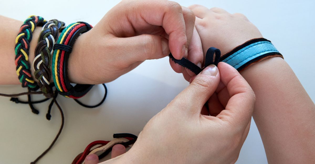 One person ties a bracelet onto another person’s wrist, with several colorful bands already worn on the arm.