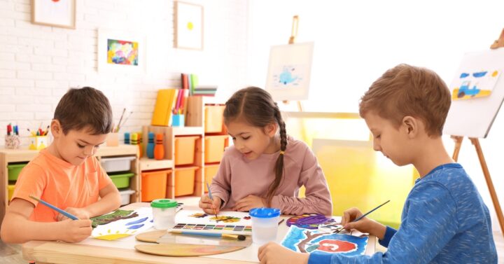 Two boys and a girl sitting around a small wooden table painting with watercolors in a room with coloring supplies.