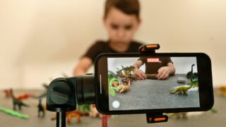 A smartphone on a tripod records a boy arranging toy dinosaurs on a table for a stop-motion movie in a playroom.