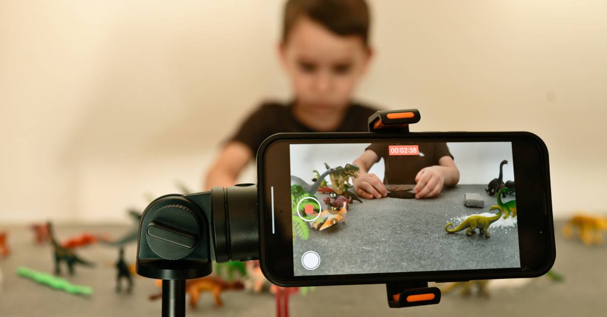 A smartphone on a tripod records a boy arranging toy dinosaurs on a table for a stop-motion movie in a playroom.