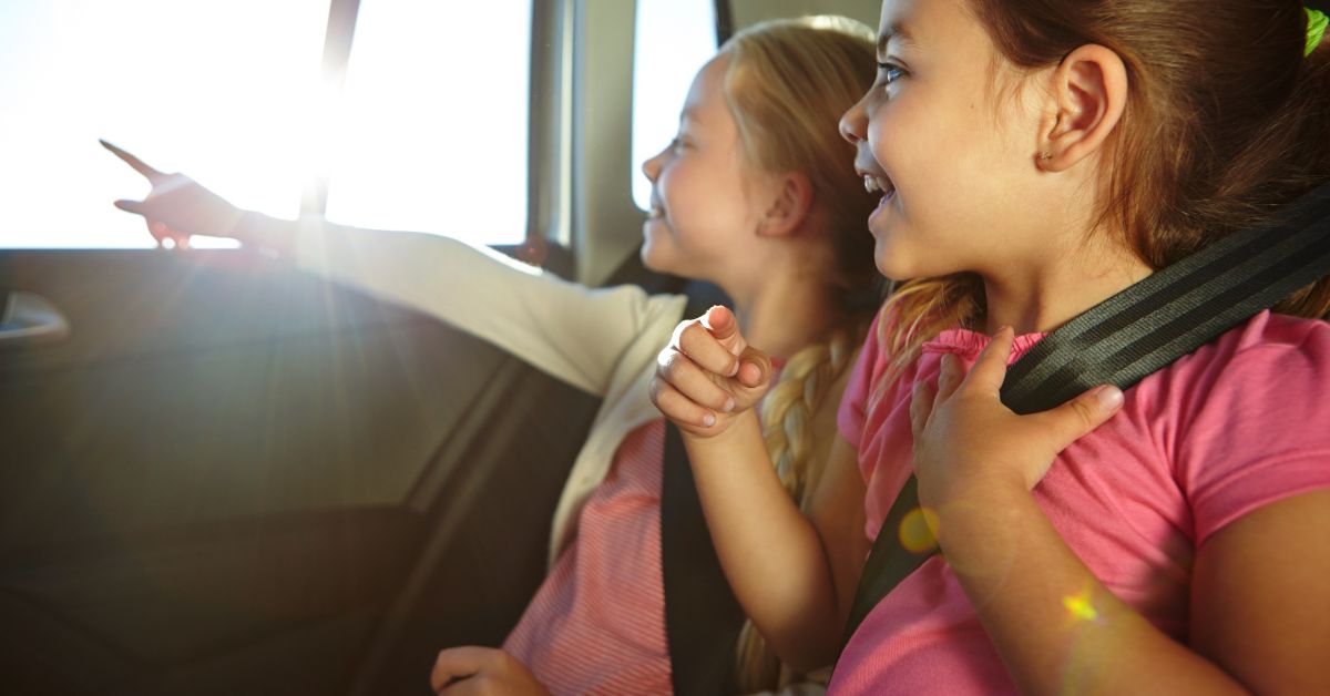 Two girls ride in the back seat of a car as one points out the window and the other smiles while holding their seat belt.