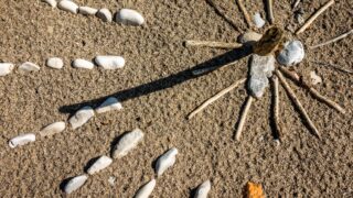 A rock-and-stick sundial casts a long shadow across the sand toward pebble hour markers in the bright sun.
