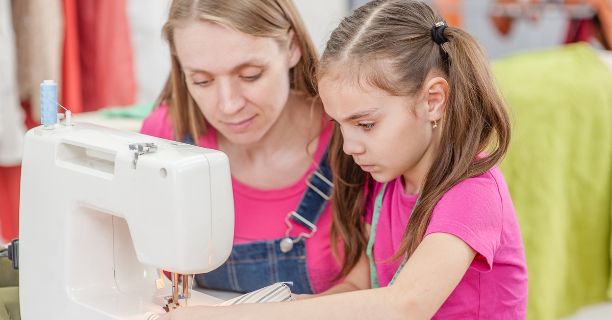 A woman and a girl wearing pink t-shirts sitting behind a sewing machine while the girl holds fabric under the needle.