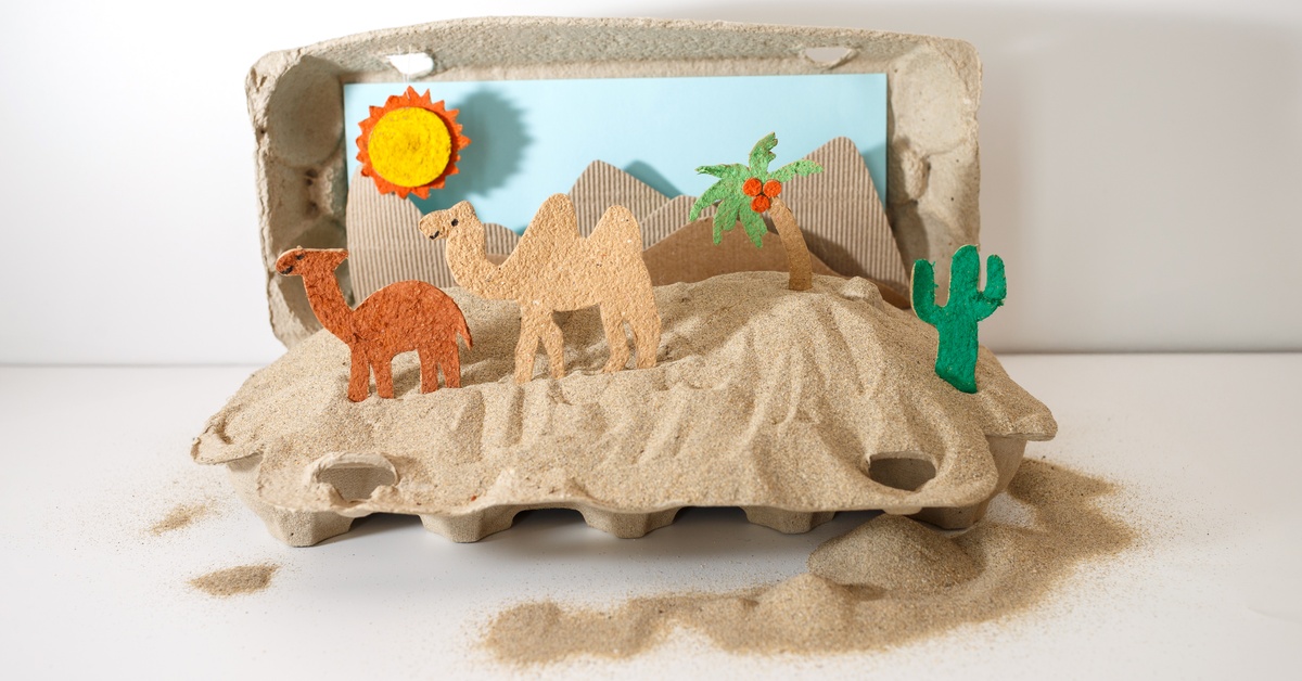 An cardboard desert diorama shows paper camels, a cactus, a palm tree, mountains, and sand under a sun.