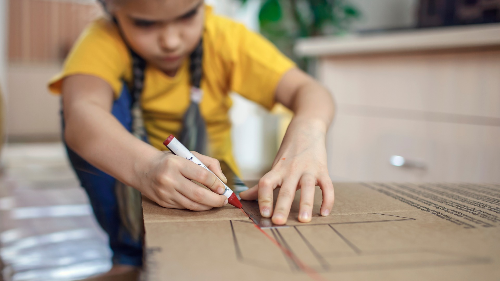 A child uses a red marker and a clear ruler to draw straight lines across a large cardboard box on the floor.
