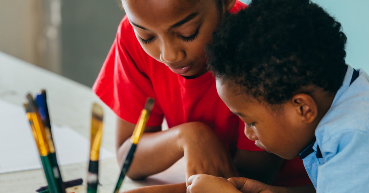 Two children leaning over a table while working on artwork, with paintbrushes and art supplies nearby.