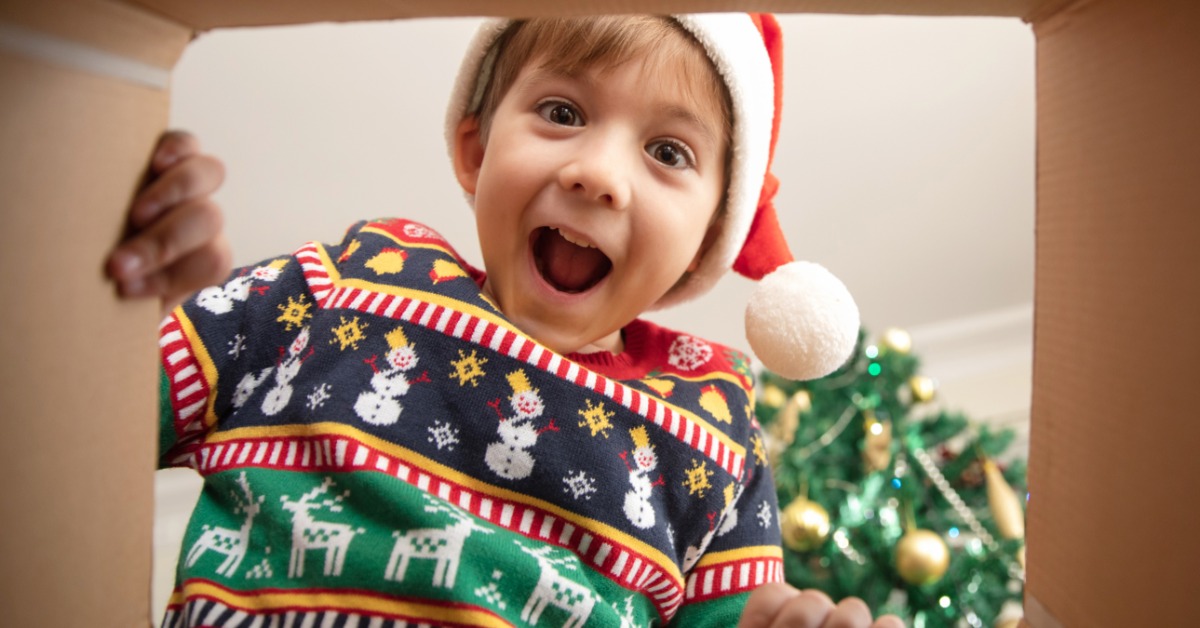 A young boy wearing a Santa hat and a Christmas sweater is looking into a cardboard box with an excited look.