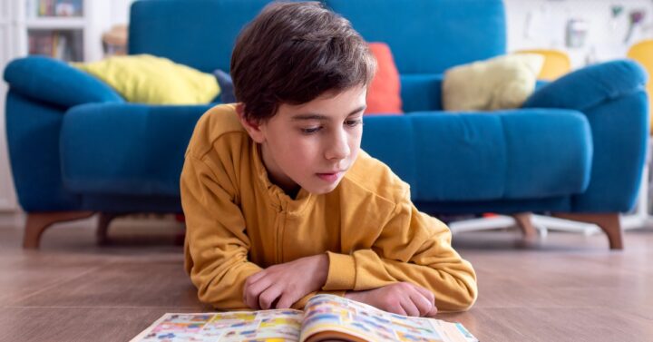 A boy lies on a living room floor and reads a colorful comic book in front of a blue sofa with bright pillows.